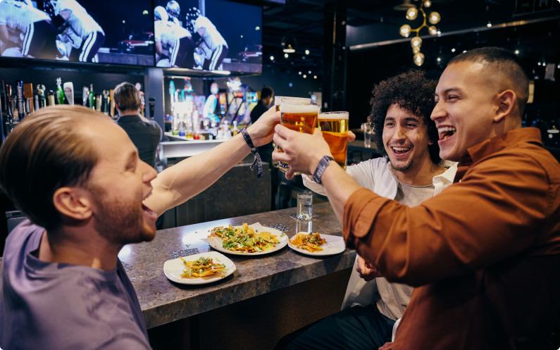 Group of three male friends toasting beers at a bar while watching sports on TV.