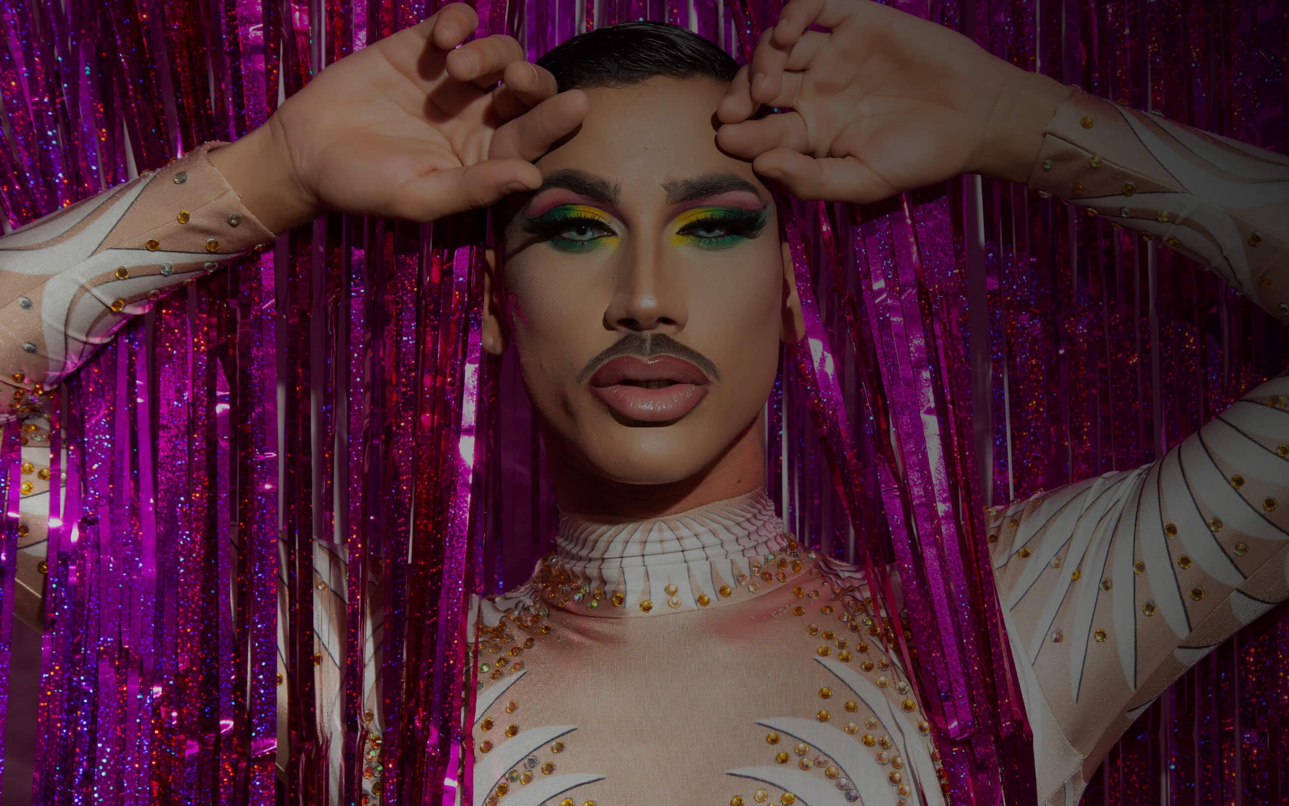 A male drag performer enters the stage through pink glittery curtains. 