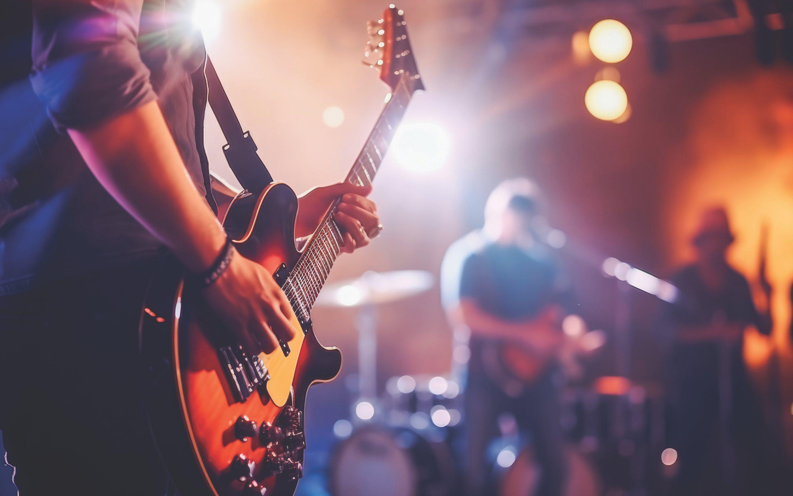 A live band performing on stage in a small venue. Performer in foreground plays a guitar. 