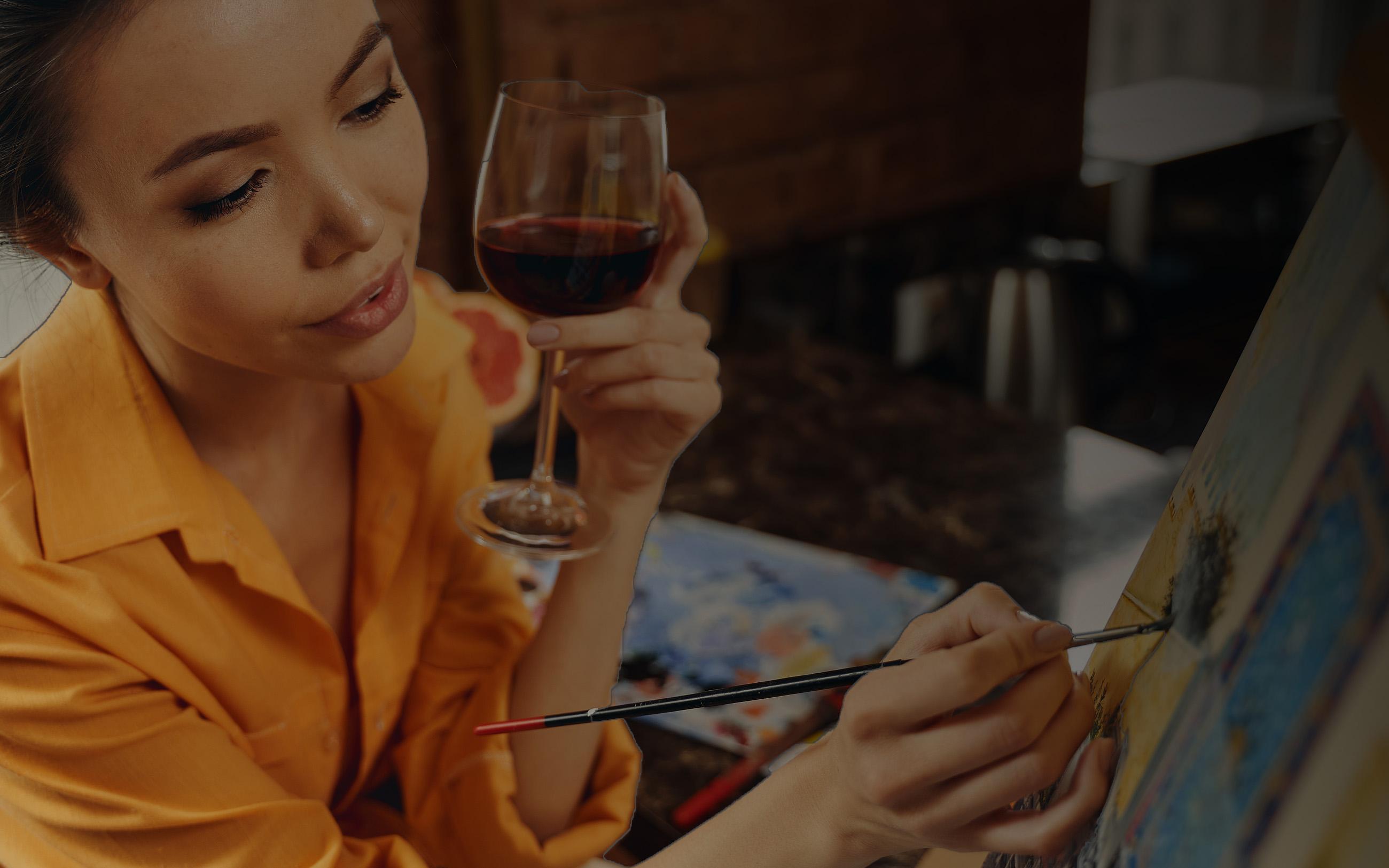 A woman enjoying a glass of wine while painting on a canvas. 