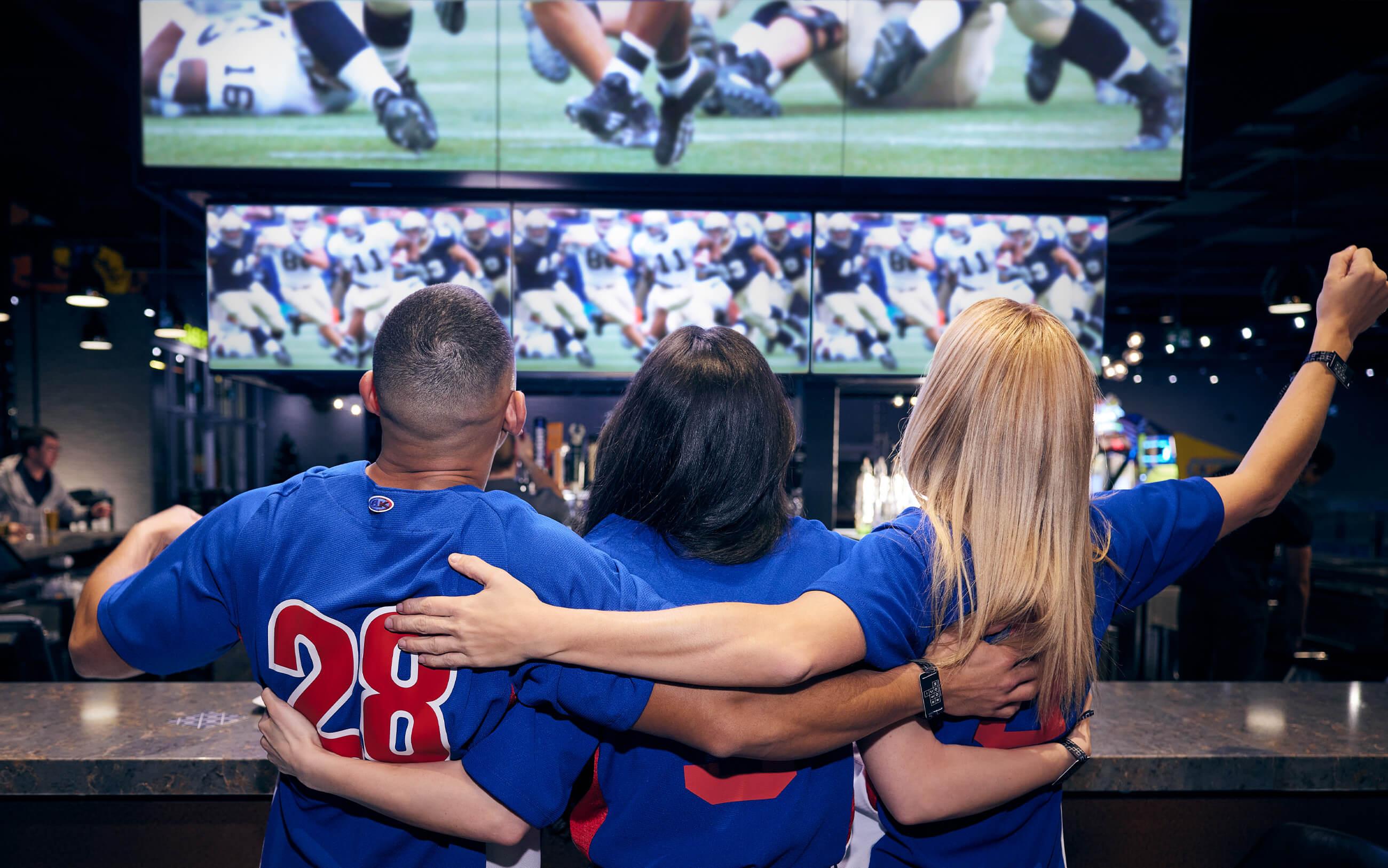 Three friends in sports jerseys celebrate while watching football on TV at the bar. 