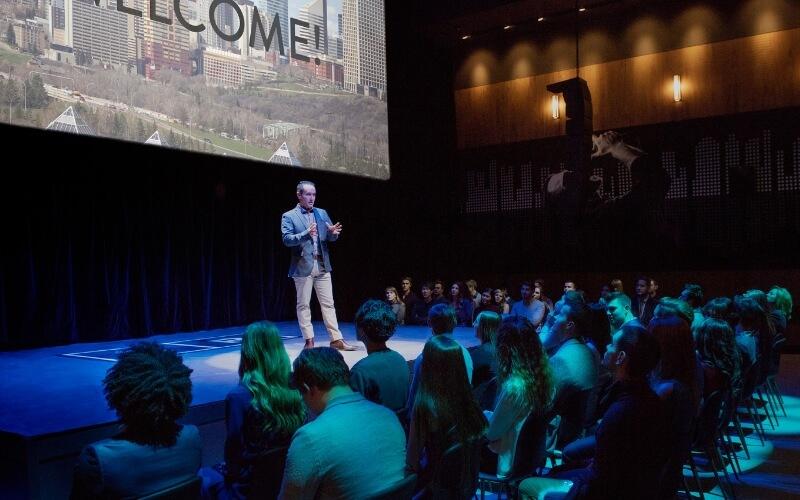 A speaker addressing an audience while standing in front of a large screen. 