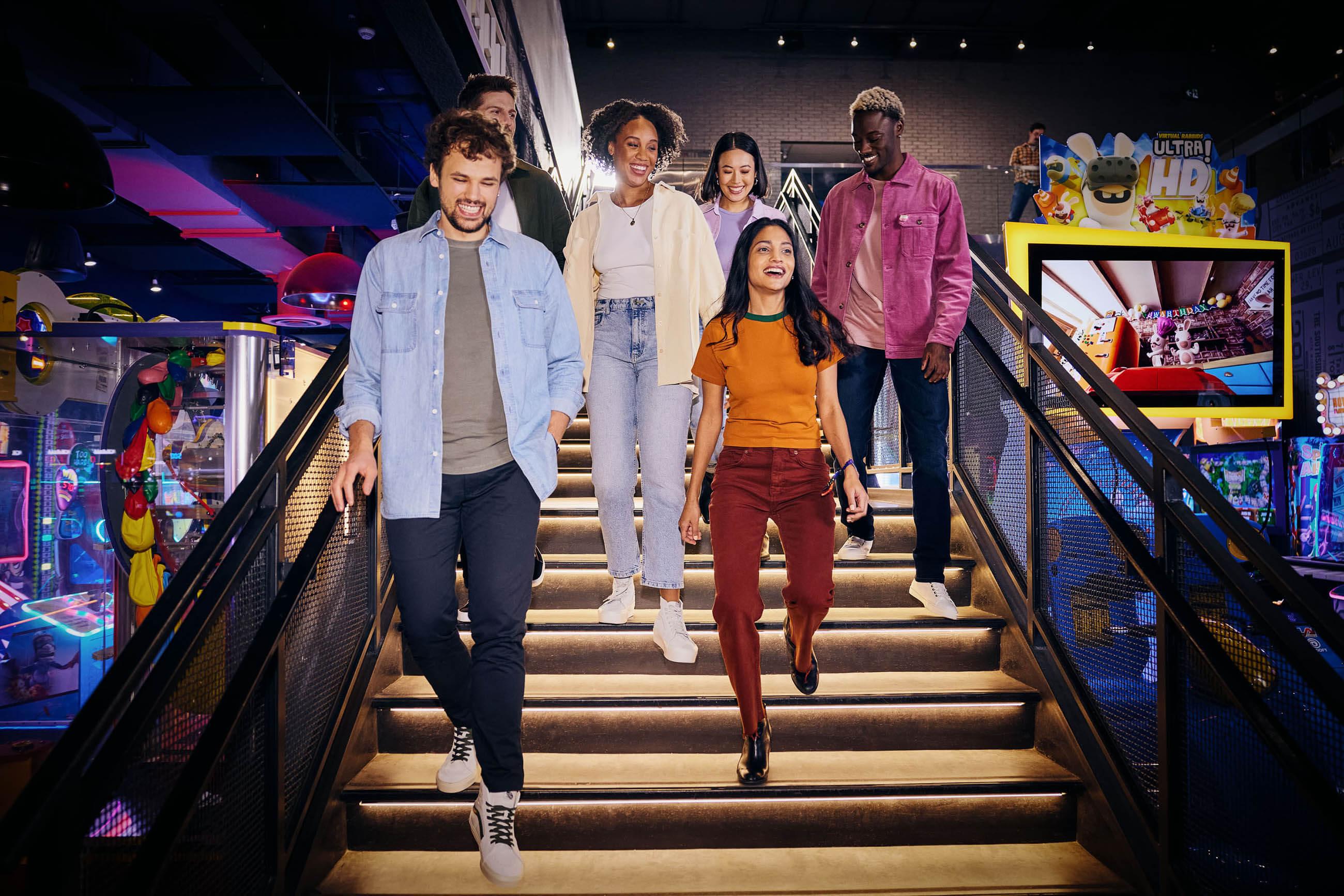 A group of 6 people coming down the stairs at The Rec Room – smiling, and ready to enjoy arcade games. 